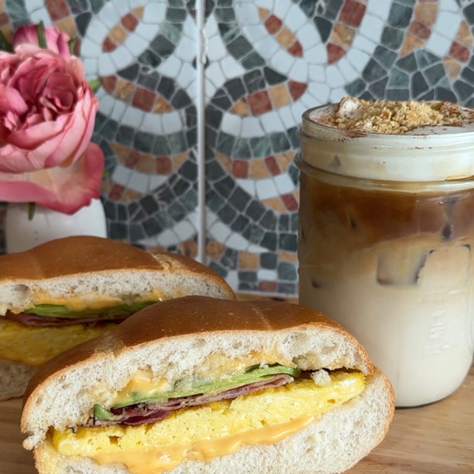 Two breakfast sandwiches with egg, cheese, bacon, and avocado, next to an iced coffee.