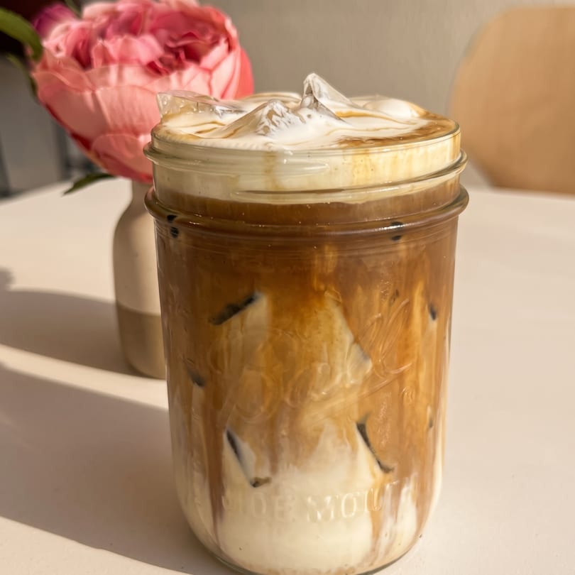 Iced coffee in a mason jar with creamy foam and caramel drizzle, a pink flower in the background.