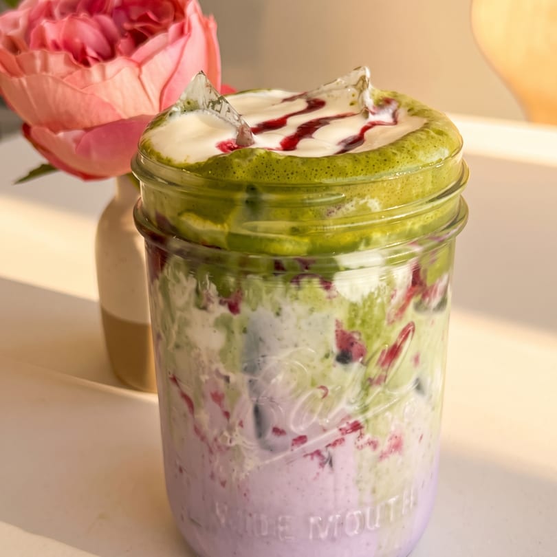 Colorful layered matcha and berry parfait in a mason jar with ice, beside a pink rose.
