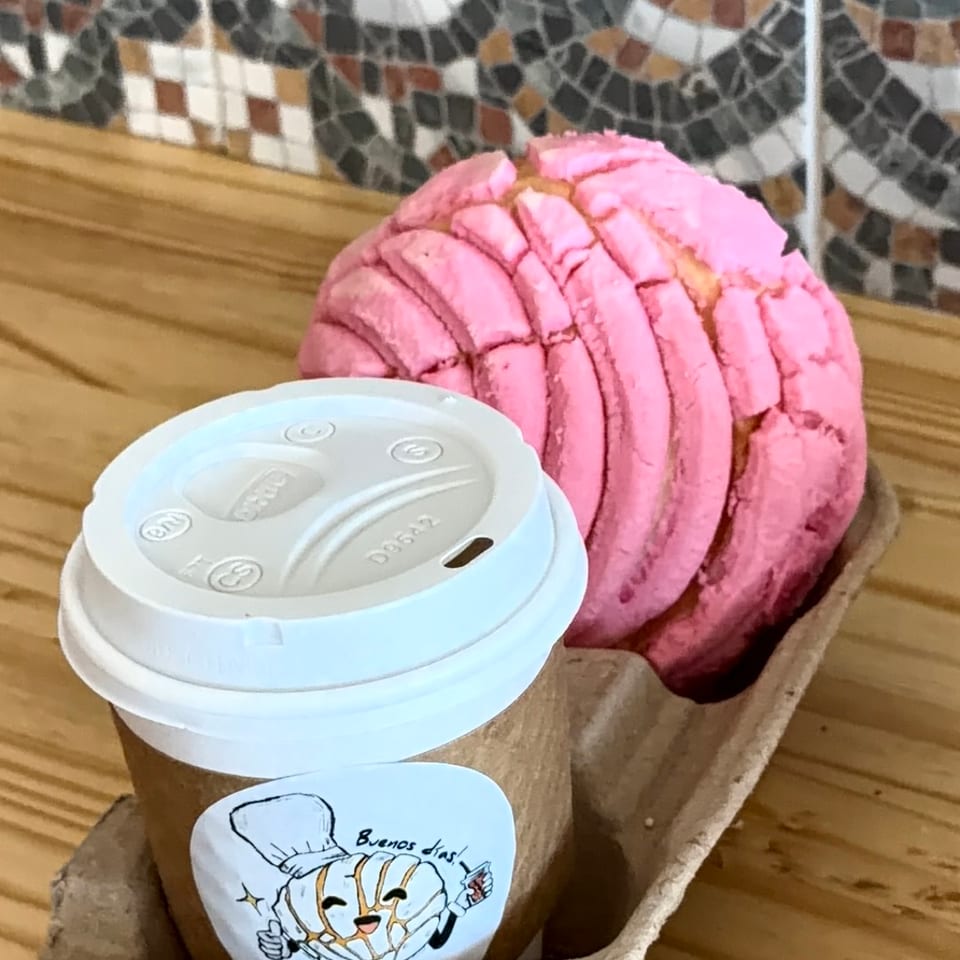 A pink concha and a coffee cup with a "Buenos dias!" sticker in a cardboard carrier.