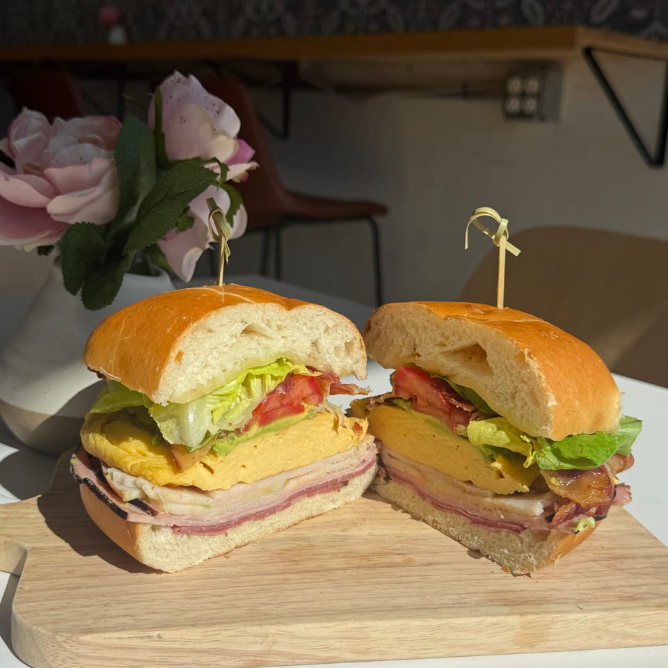 Two halves of a large sandwich with egg, deli meats, lettuce, tomato, and avocado on a cutting board.