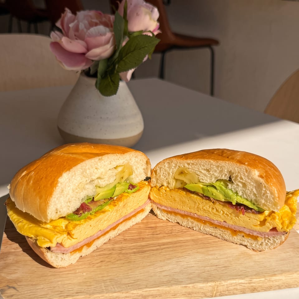 Sandwich with layered egg, ham, and avocado, sliced in half on a wooden board in sunlight.