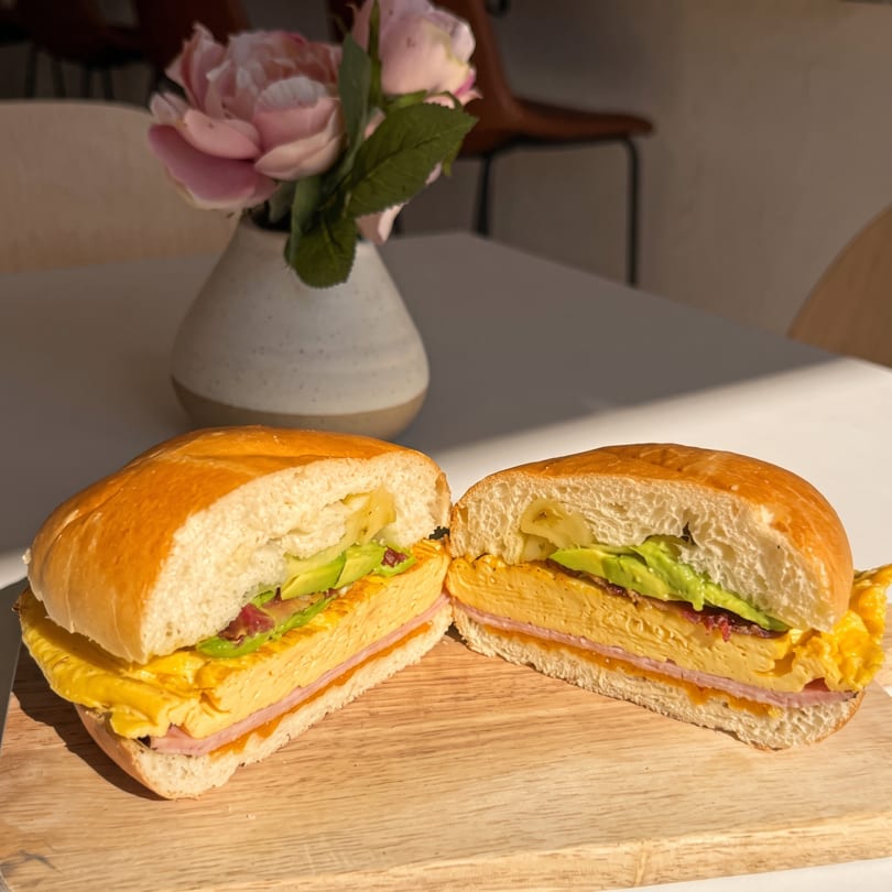 Sandwich with layered egg, ham, and avocado, sliced in half on a wooden board in sunlight.